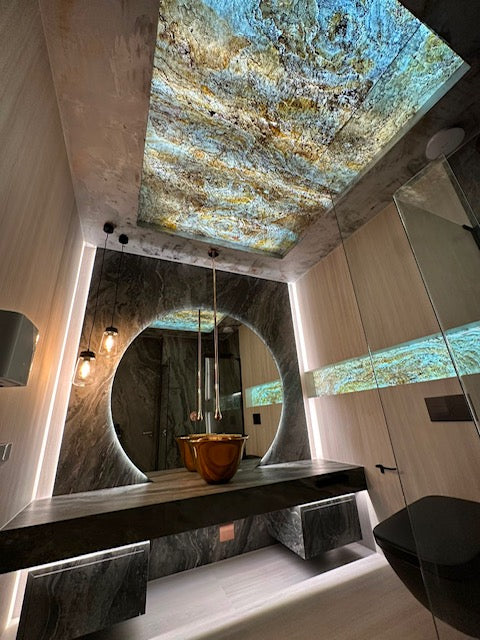 A luxury modern bathroom featuring a large circular mirror, a floating dark stone vanity, a copper vessel sink, and a dramatic light-up ceiling with a textured blue and gold pattern.